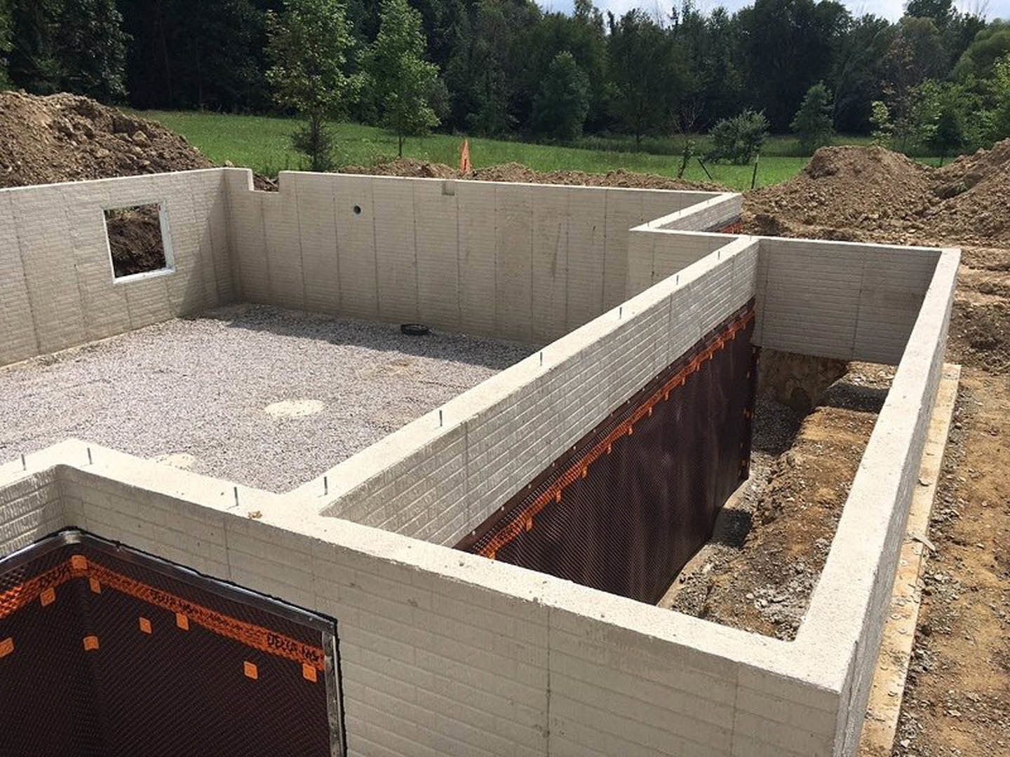 Concrete foundation and partially built exterior wall surrounded by temporary fencing, with open field and trees in the background.