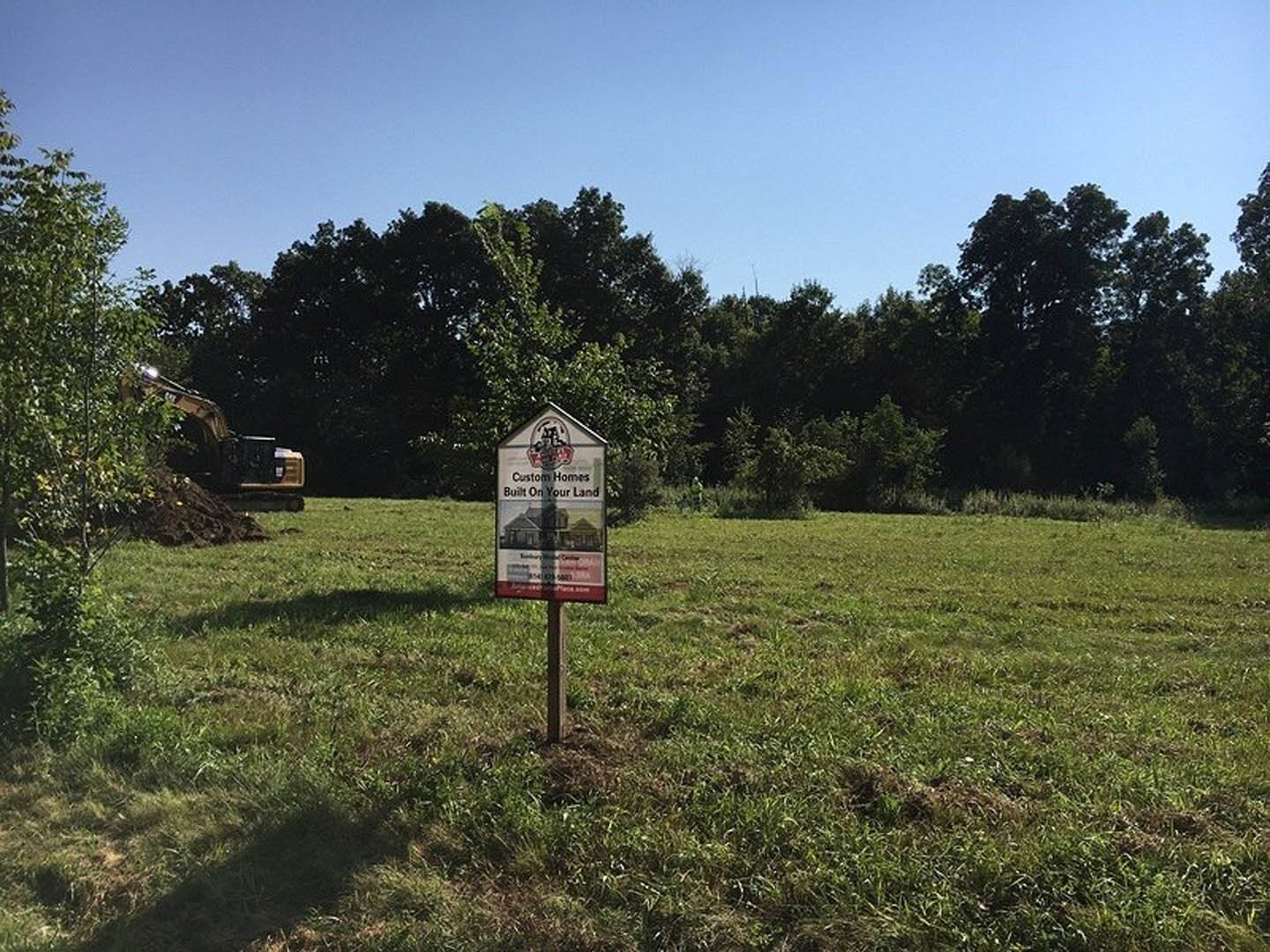Wooden post sign marking a grassy field with scattered trees under clear blue sky, land lot prepared for residential construction.