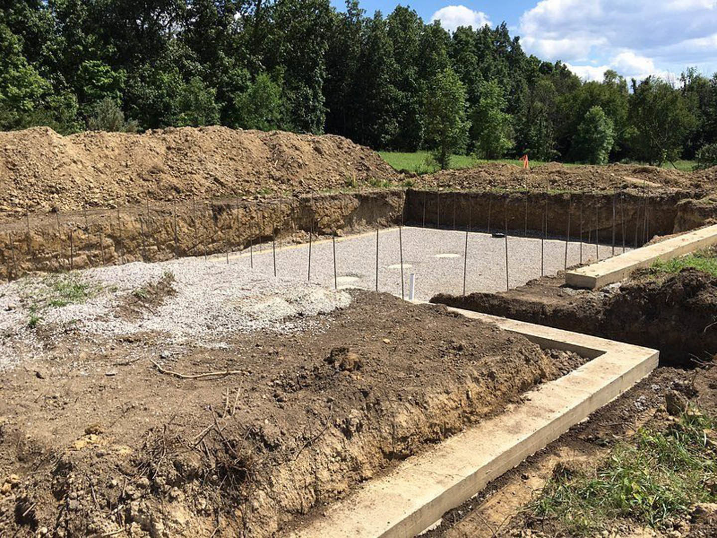 Concrete foundation forms set in a dirt excavation surrounded by soil mounds, rocks, and scattered wooden stakes, with trees and blue sky dotted with white clouds in the background