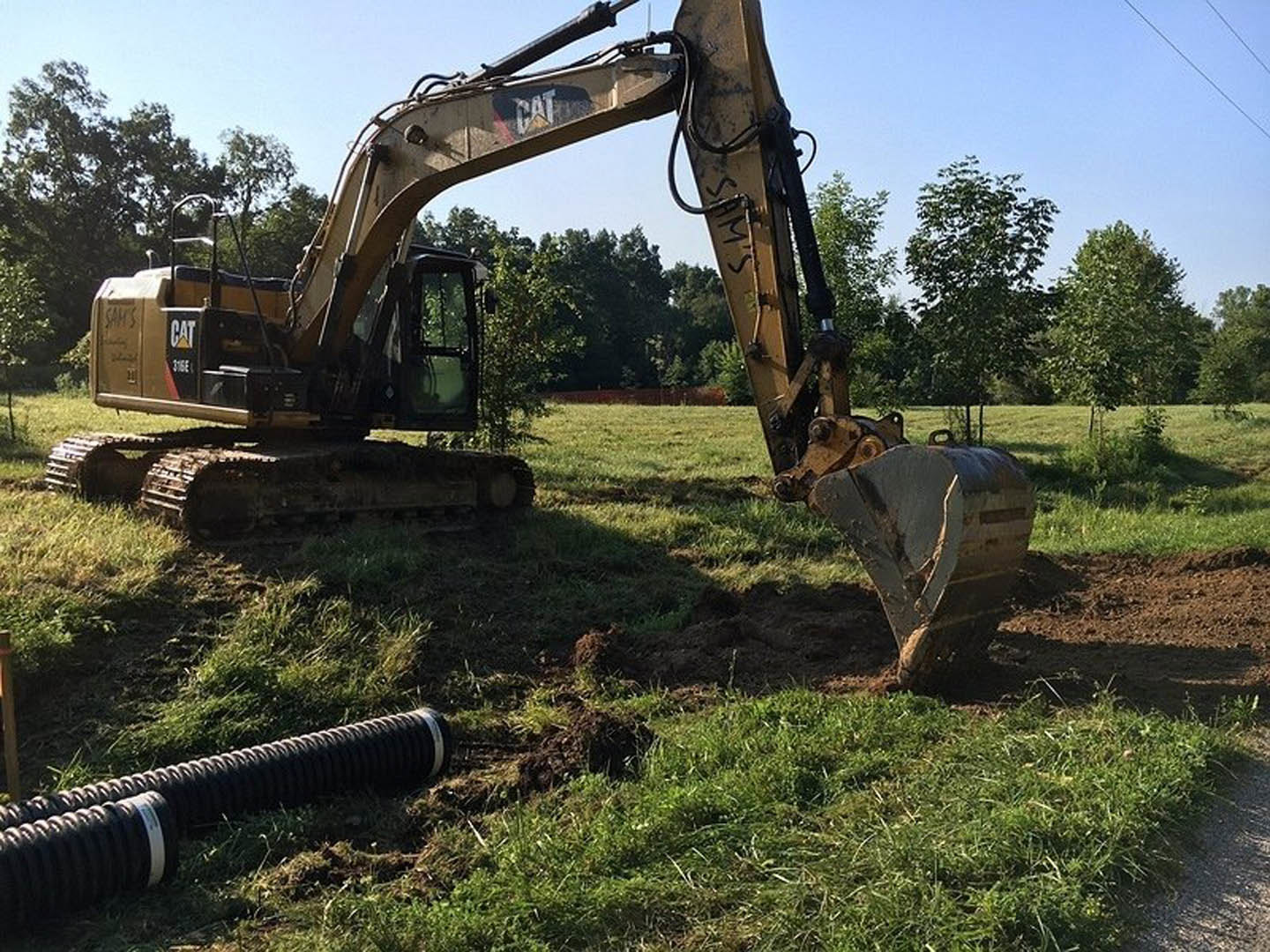 Yellow bulldozer parked on grassy field beside black drainage pipe, blue sky and trees in background