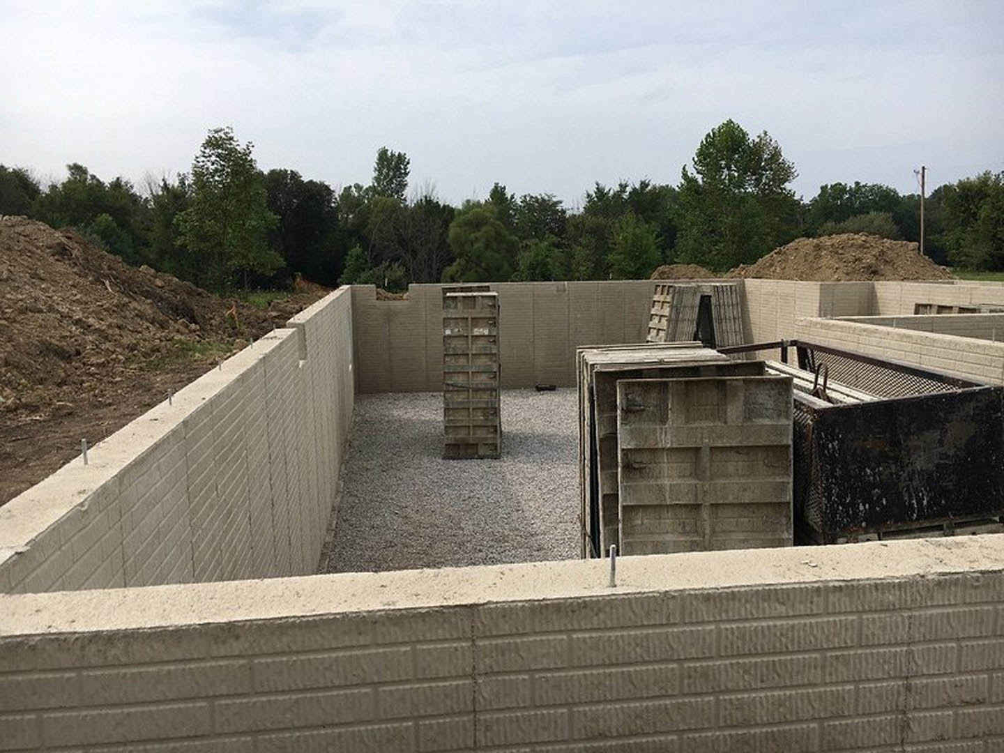 Concrete foundation with a pile of cement, adjacent concrete wall, wooden door with metal handle, black metal box, and pallets on the ground; outdoor setting with trees and cloudy
