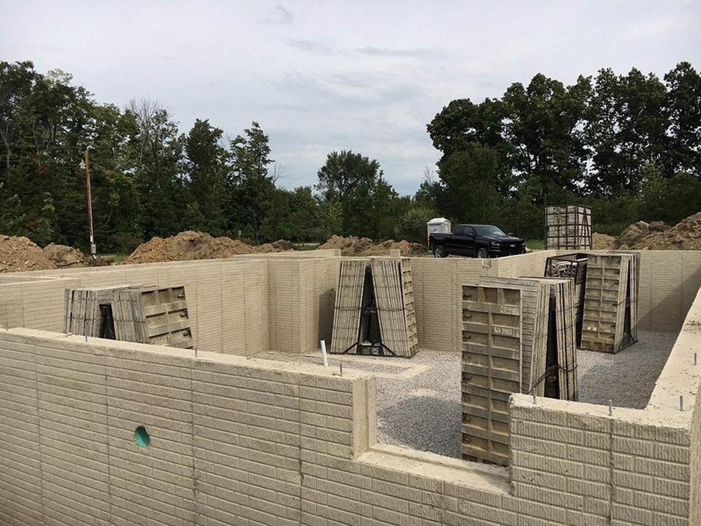 Modern home exterior with triangular roof, large window, and prominent concrete wall; black truck parked roadside; mature trees and overcast sky in background.
