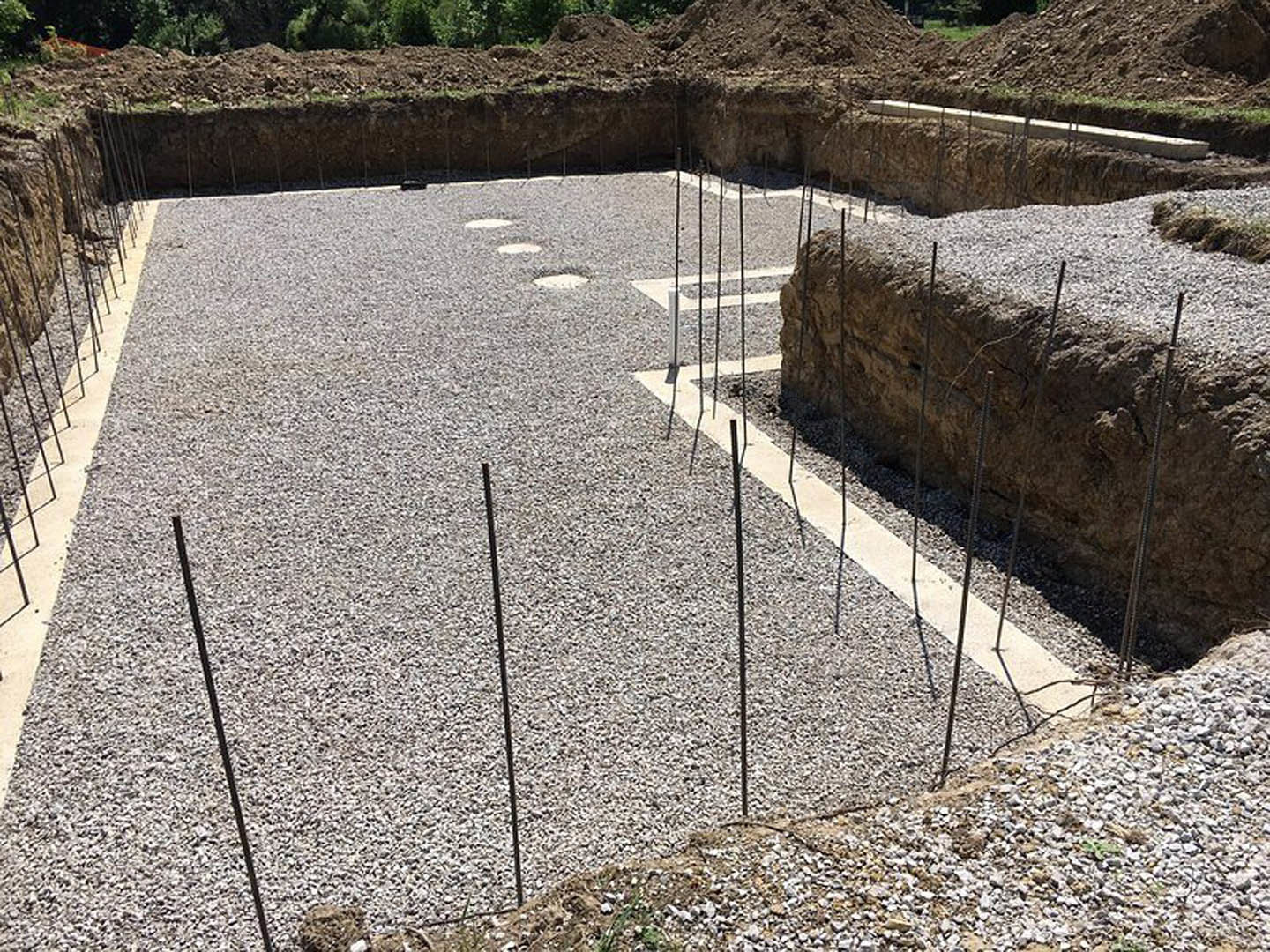 Gravel foundation with embedded metal rods, surrounded by construction materials and unfinished ground.