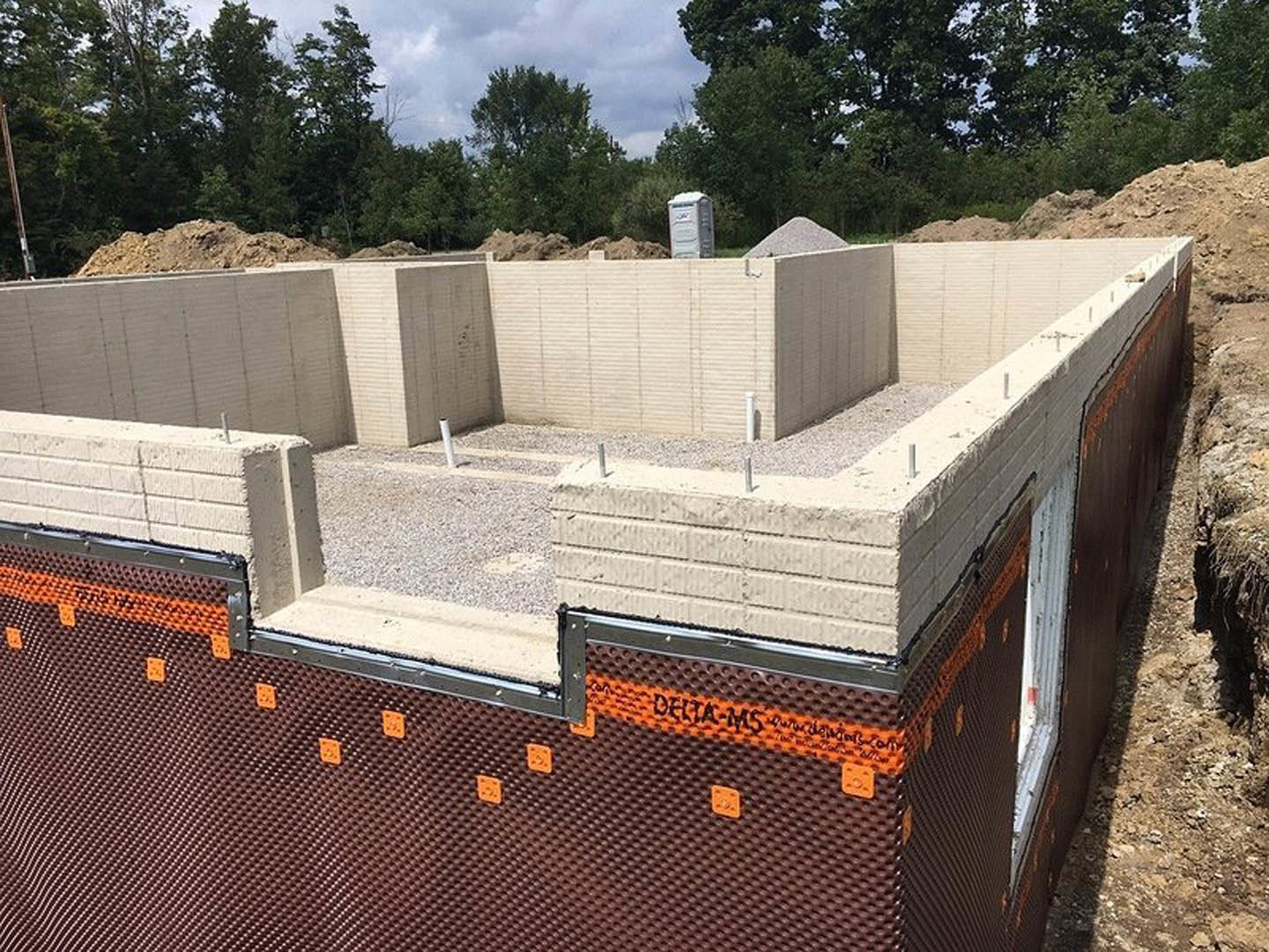 Partially built home with exposed concrete foundation, white construction materials, piles of dirt, and trees in the background