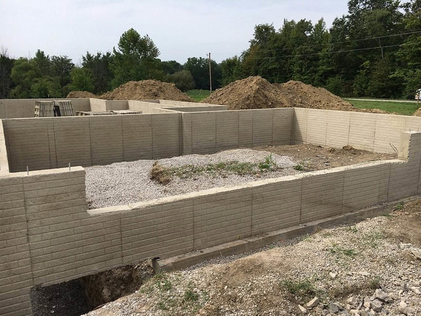 Concrete foundation surrounded by dirt and grass, brick wall section, pile of soil, and trees in the background under a clear sky