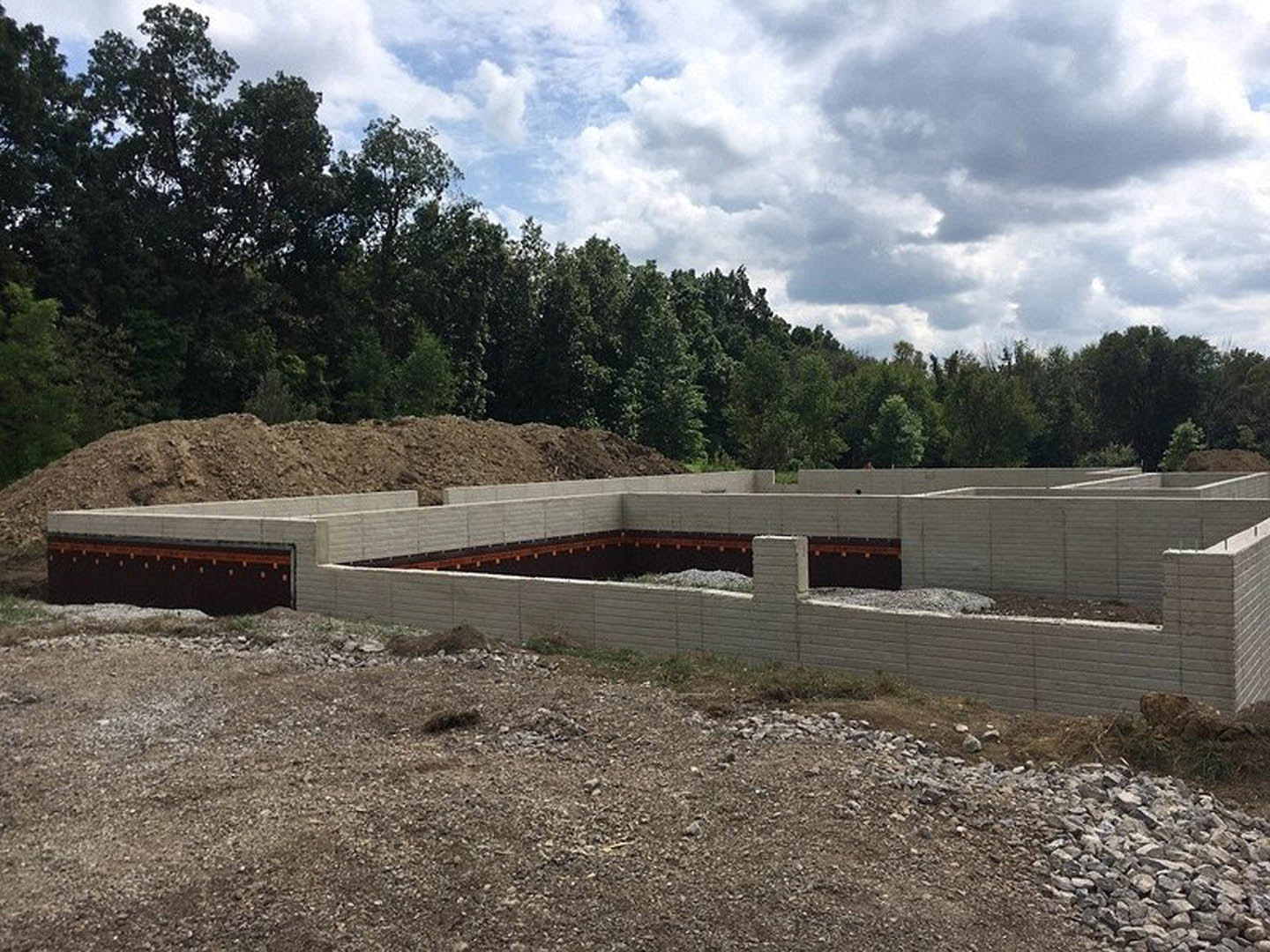 Concrete foundation wall with a circular opening, surrounded by dirt, rocks, and a pile of stones, under a cloudy sky with trees in the background