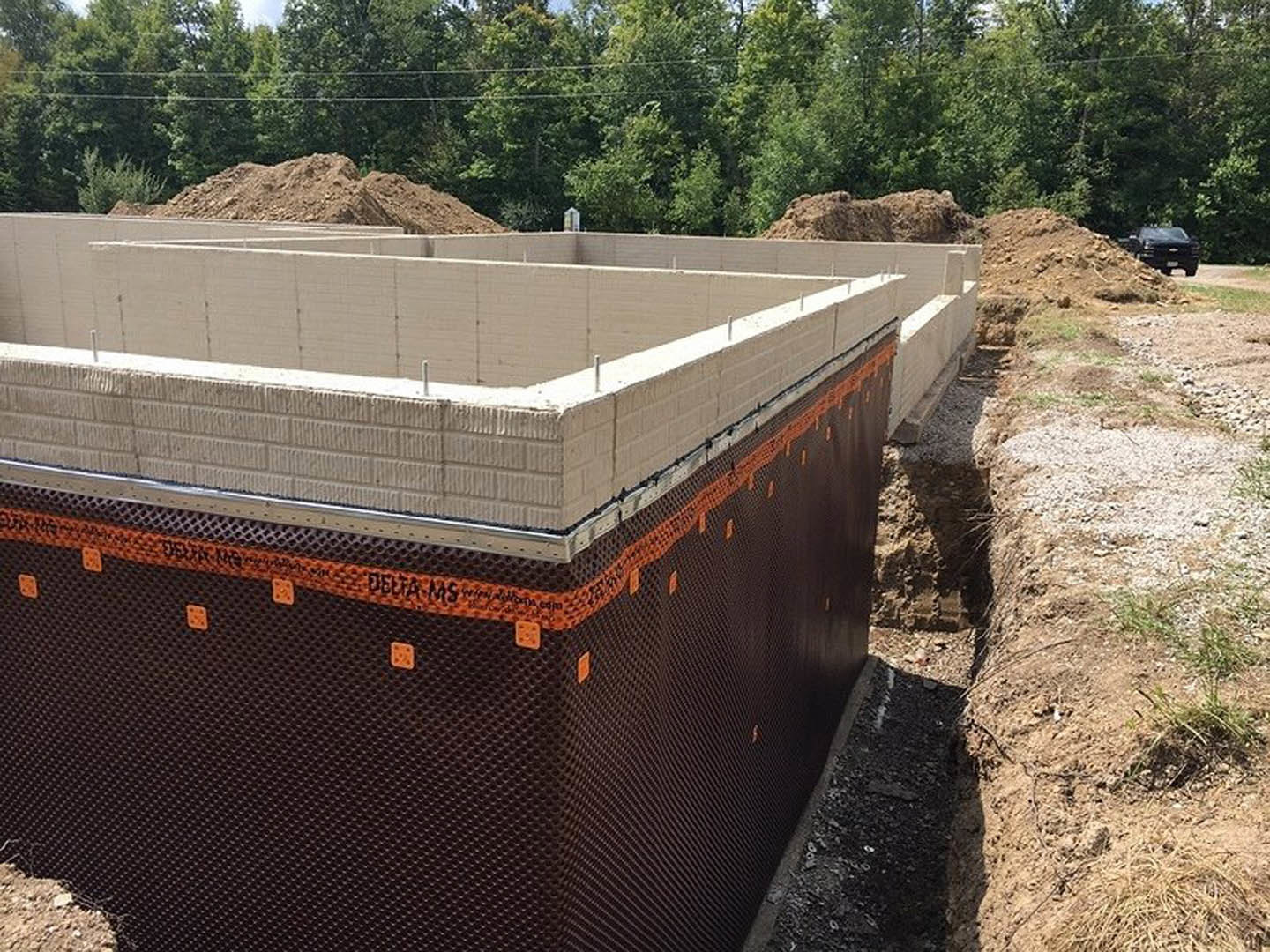 Concrete foundation with partially built block wall, surrounded by dirt, grass, and nearby trees with visible power lines in the background