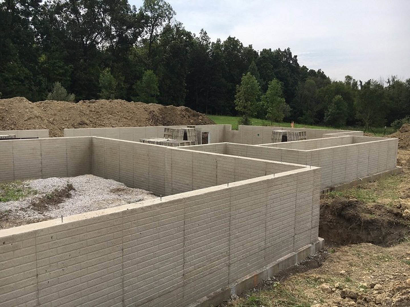 Partially constructed foundation walls with a central opening, piles of dirt and gravel on the ground, scattered construction materials, dirt hill and trees in the background under