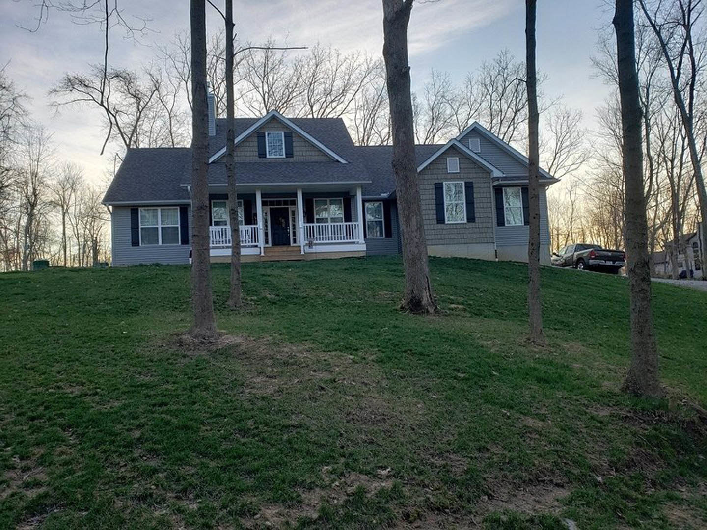 Two-story house with white porch railing, surrounded by trees on a grassy hill, black truck parked in driveway, close-up view of front door and windows