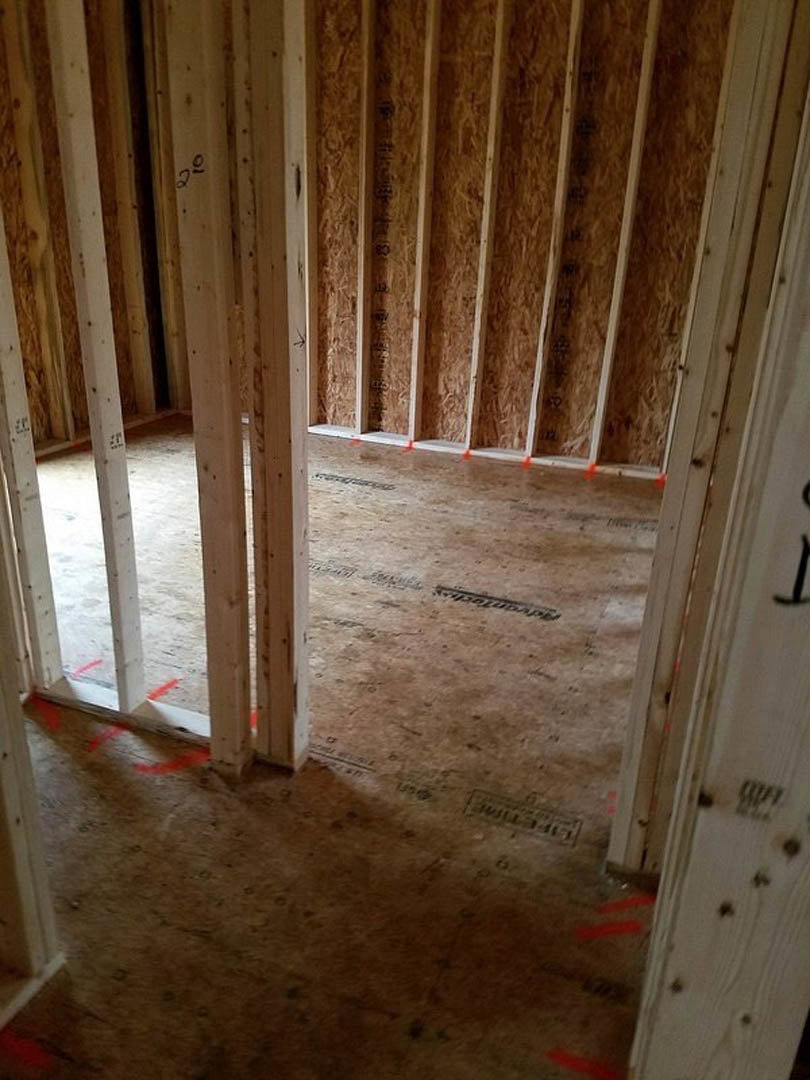 Unfinished room with exposed wood framing, two doorways, subfloor planks, wall insulation, and construction materials scattered on the floor