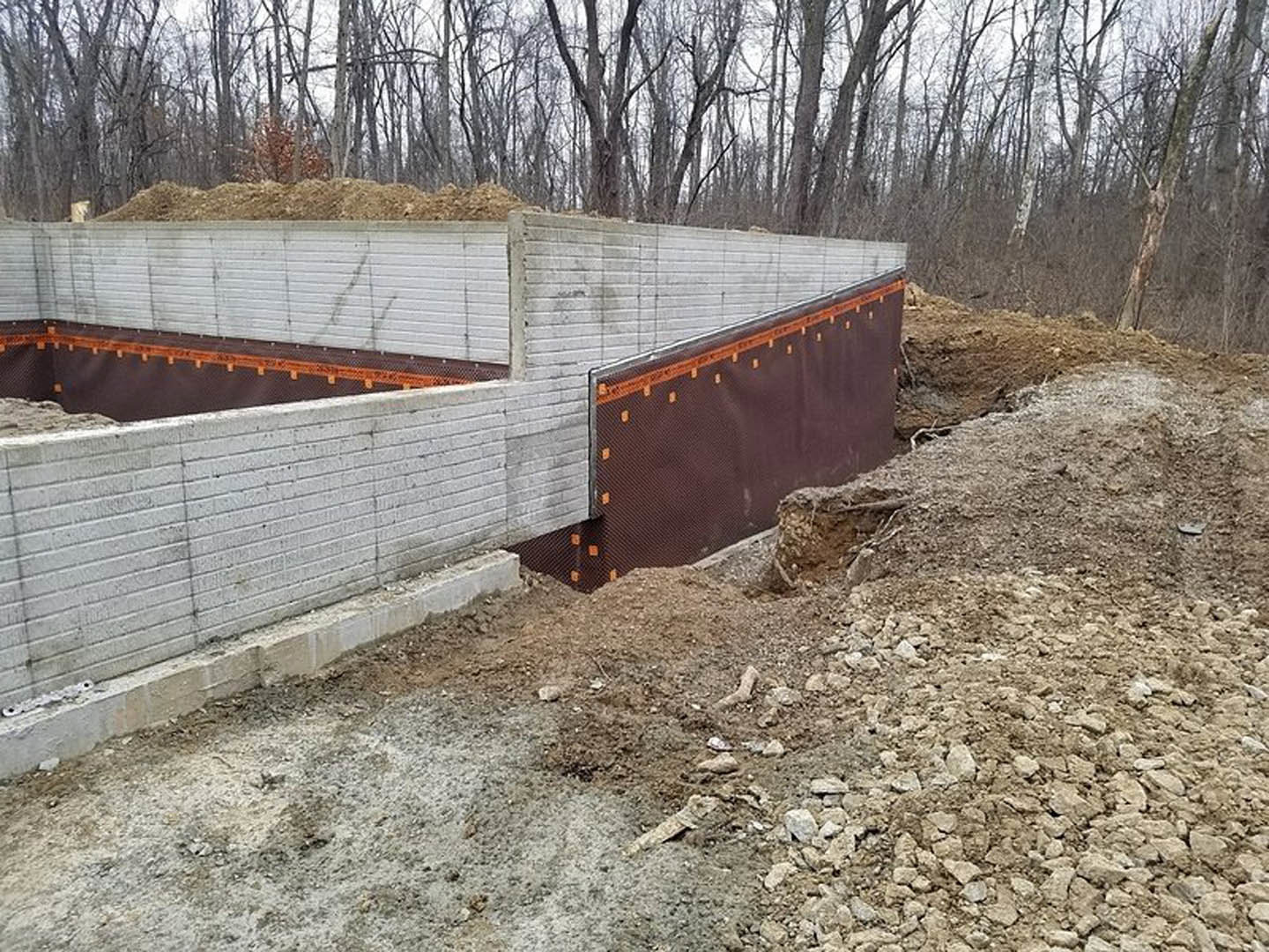 Concrete foundation wall with exposed dirt and rocks, tree adjacent to wall, open hole in ground, outdoor construction site with fence and sky visible