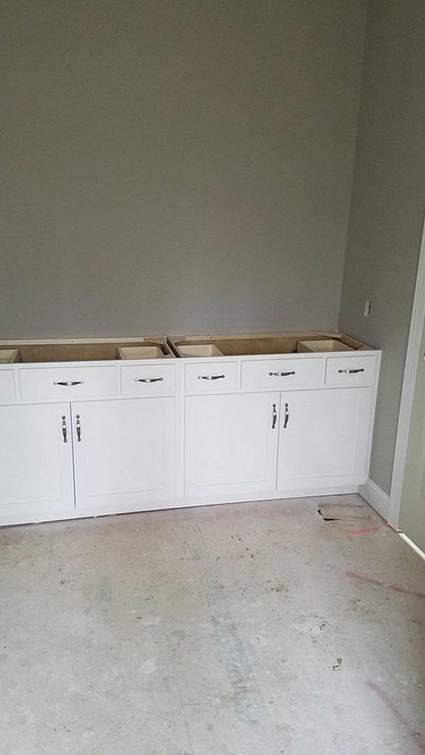 White kitchen cabinetry with flat-panel doors, light wood flooring, plaster walls, and empty countertops in a modern, minimalist room.