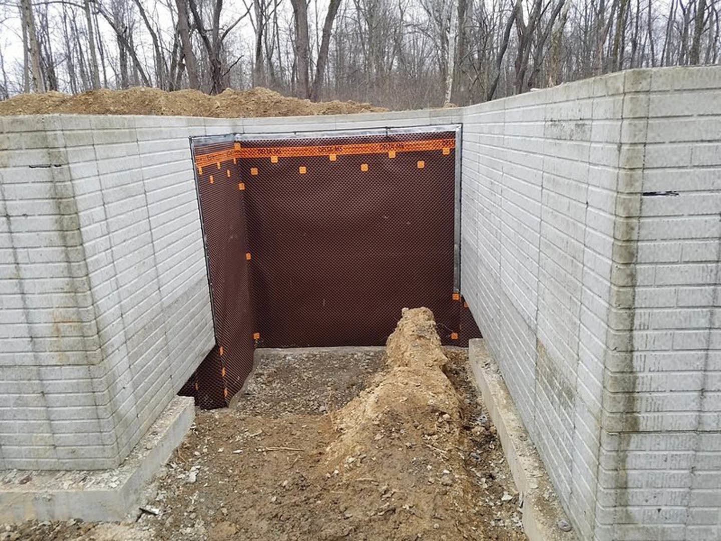 Smooth concrete wall with circular hole, surrounded by dirt and rocks, brick wall with red and black mesh in background, group of trees visible beyond exterior structure