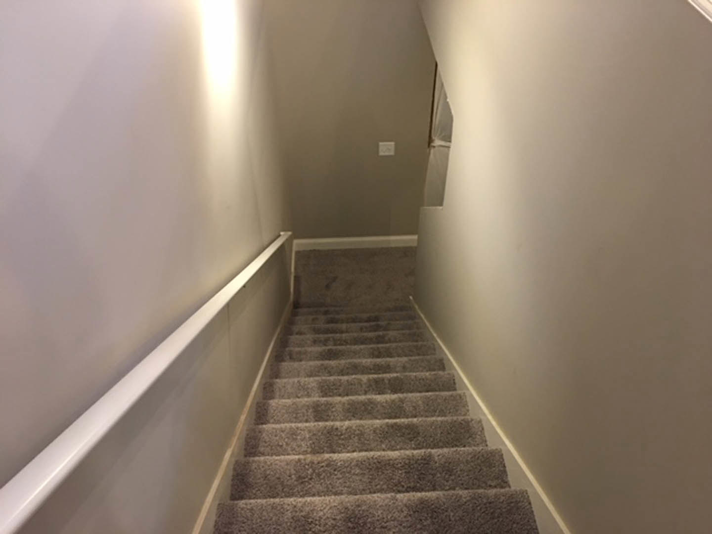 Carpeted staircase with white plaster walls, wall-mounted light fixture, white door with matching handle, and visible light switch