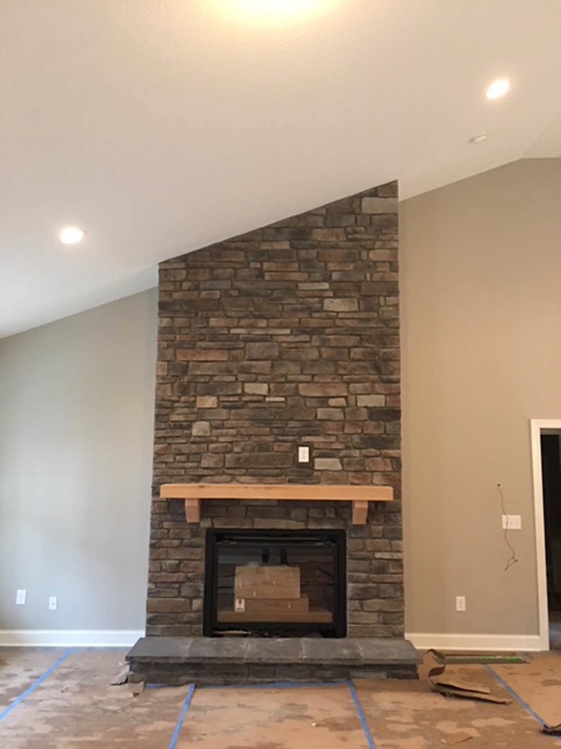 Brick fireplace with wood mantel, stacked firewood boxes, stone accent wall, hardwood flooring, and fire screen in a cozy living room.