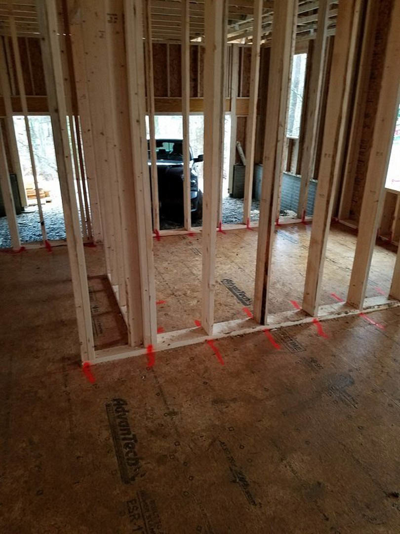 Wood-framed room under construction with exposed beams, red tape on the subfloor, and a car visible through the window in the background