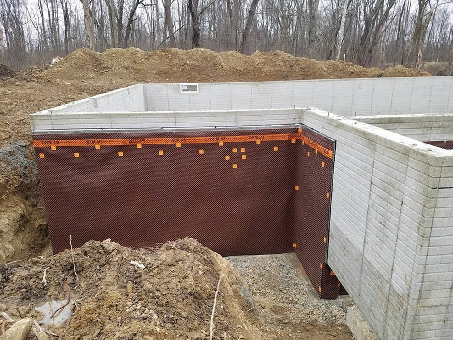 Concrete foundation slab surrounded by dirt piles and construction materials, with trees and blue sky in the background