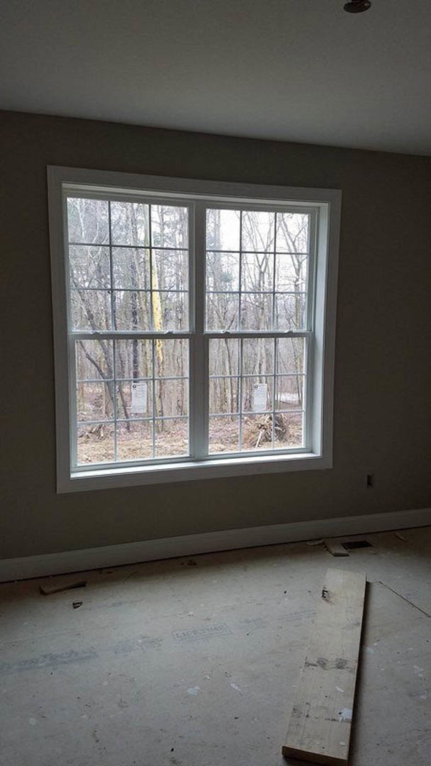 Large window with white blinds set in plaster wall, natural daylight streaming onto light wood floor, leafy trees visible outside