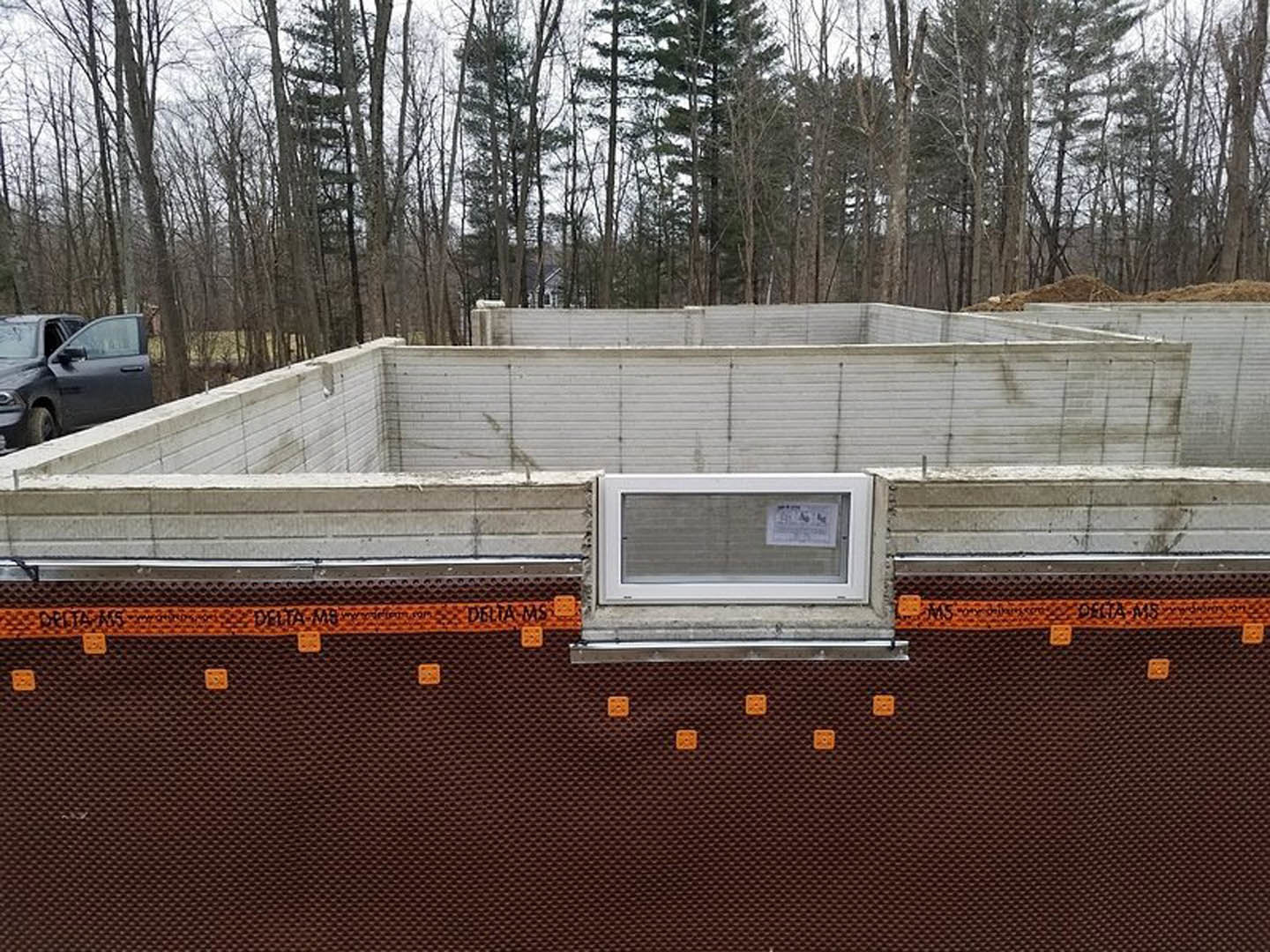 Partially built house framed with exposed wood, construction materials scattered around, black pickup truck parked nearby, temporary fencing and trees in background, multiple