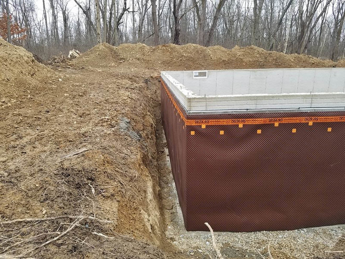 Concrete foundation slab surrounded by dirt piles and brown tarp, set in a wooded area with trees and grass, black wire visible along white wall, rectangular object placed in