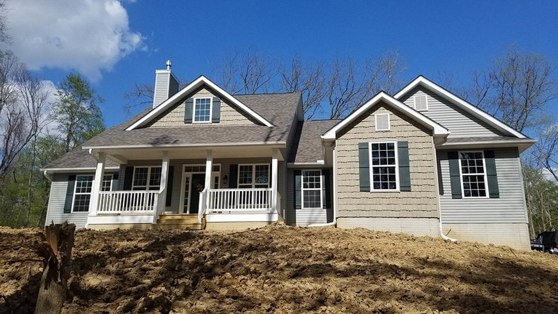 Two-story house with light siding, covered front porch, and windows, set beside a dirt hill and surrounded by trees