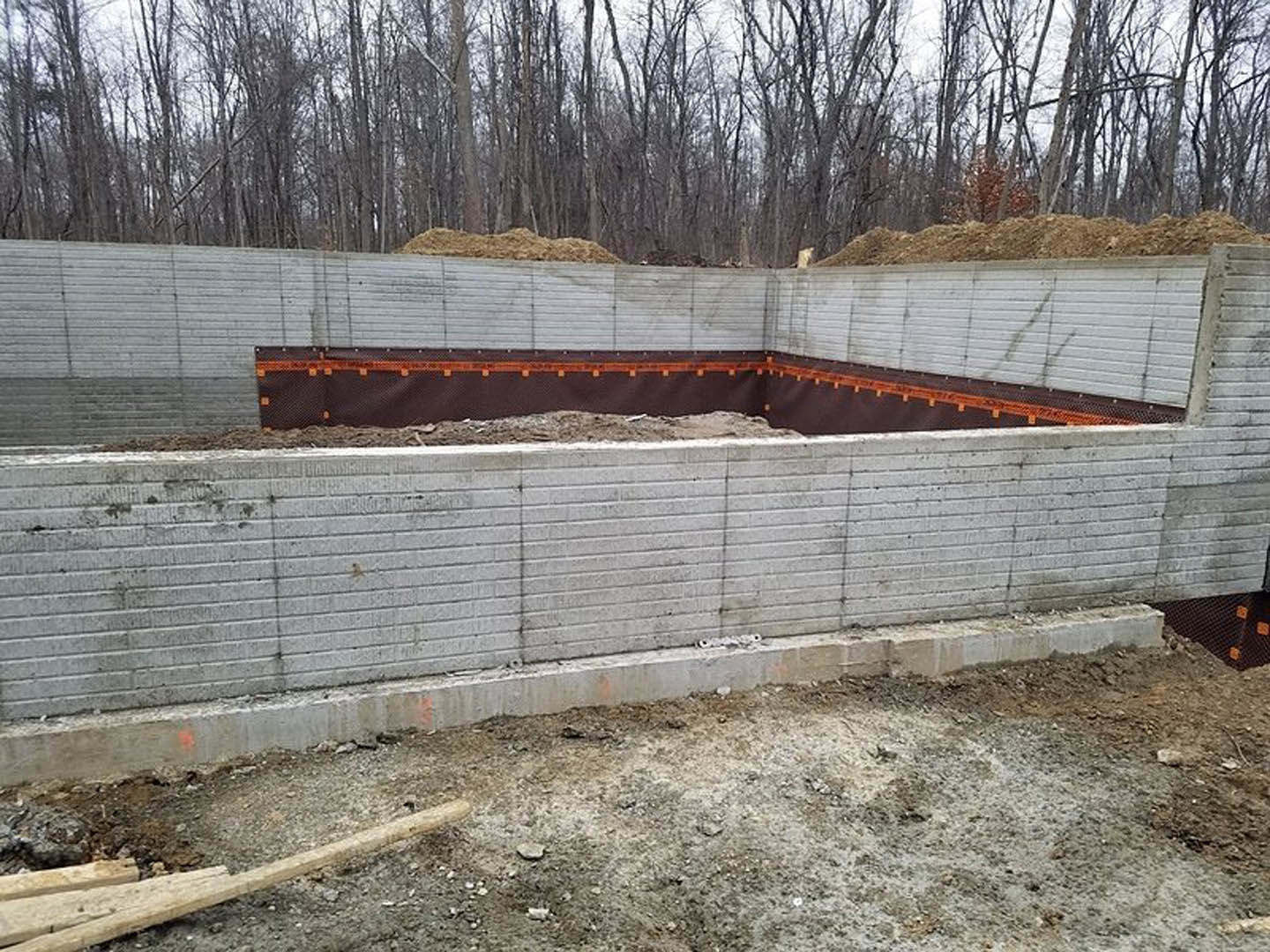 Concrete foundation wall surrounded by dirt and rocks, wooden plank lying on ground, trees in background.
