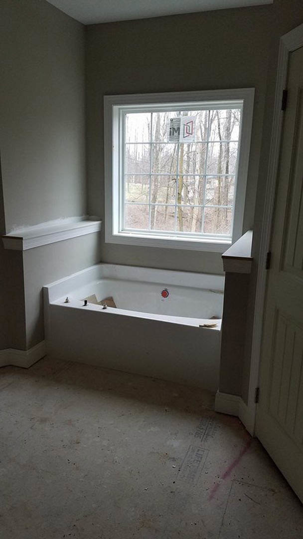 Freestanding white bathtub on light wood floor beside large window, plaster walls, and minimalist fixtures in modern bathroom