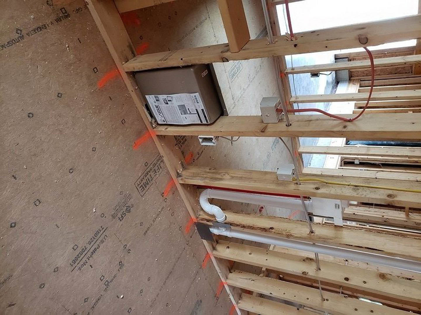 Exposed wood framing with electrical wires, plumbing pipes, a labeled electrical box, and orange tape marking on a wood shelf in a residential construction interior