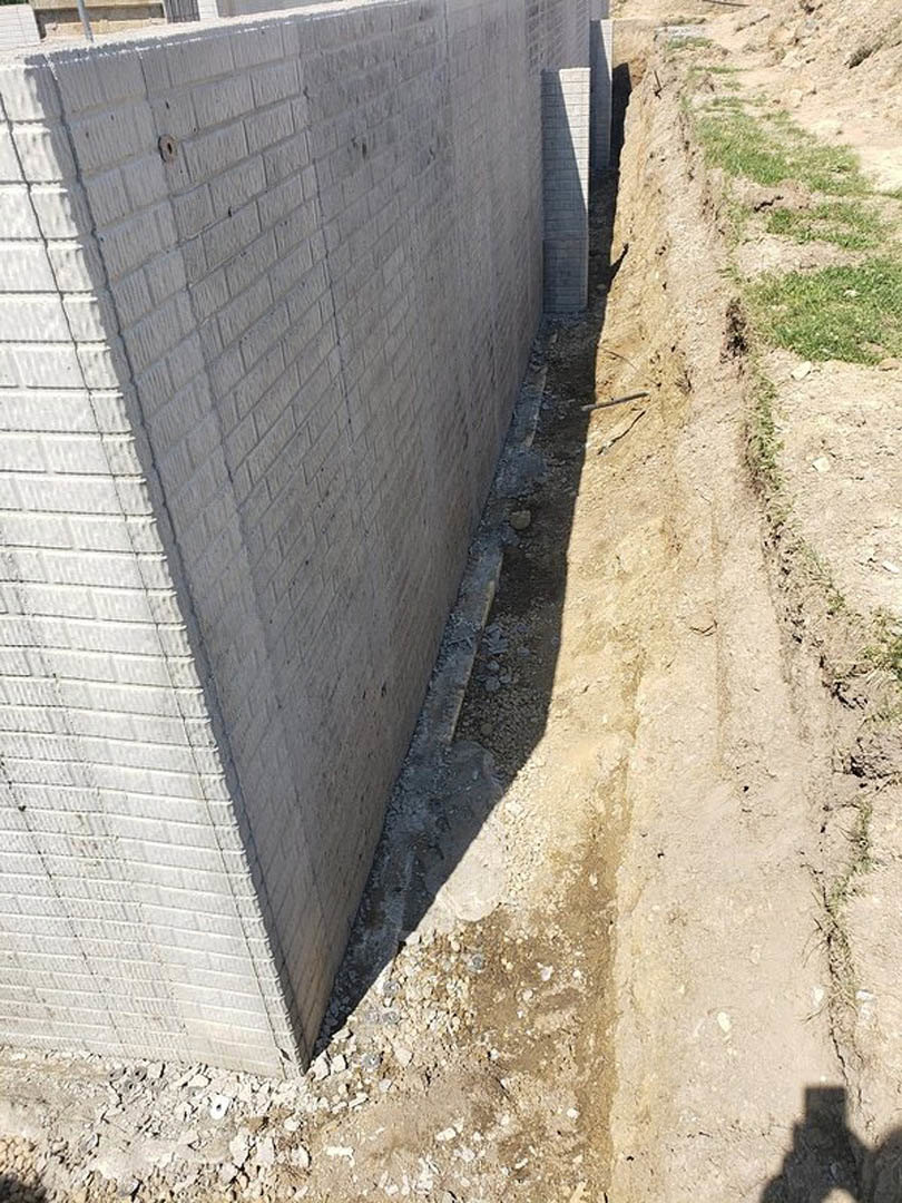 White brick exterior wall with a rough cement base, dirt patch and hole in the ground, shadow of a person cast across the outdoor surface.