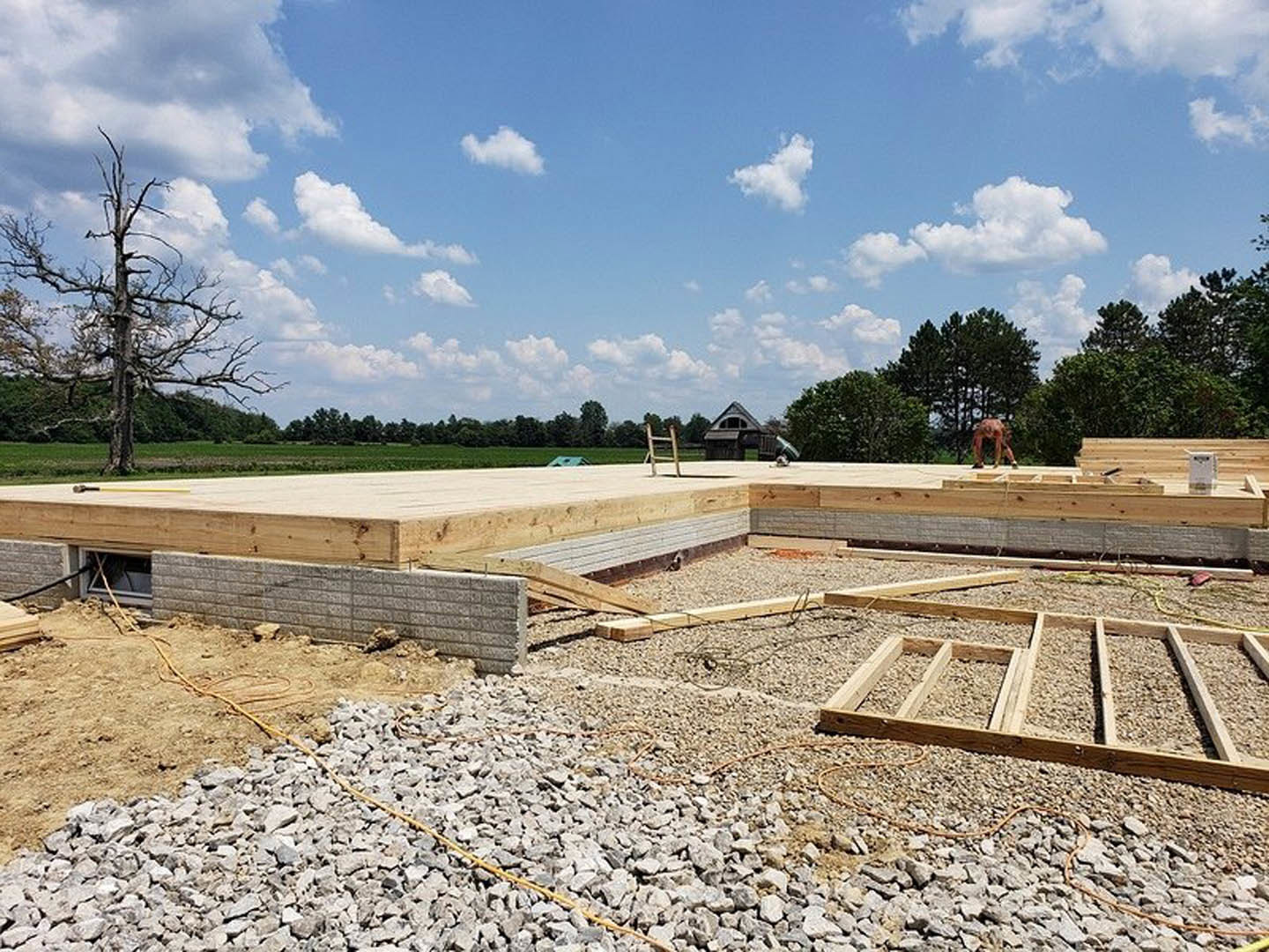 Wooden framing structure on gravel ground, person working nearby, leafless tree, blue sky with clouds, pile of rocks and wooden beam in foreground