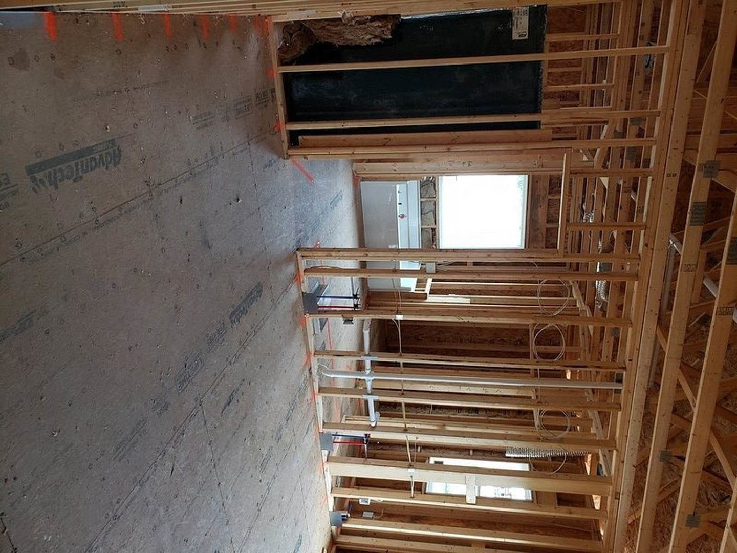 Exposed wooden framing and beams inside a house under construction, unfinished walls, window with light visible in background, white door with glass panel, lumber and construction
