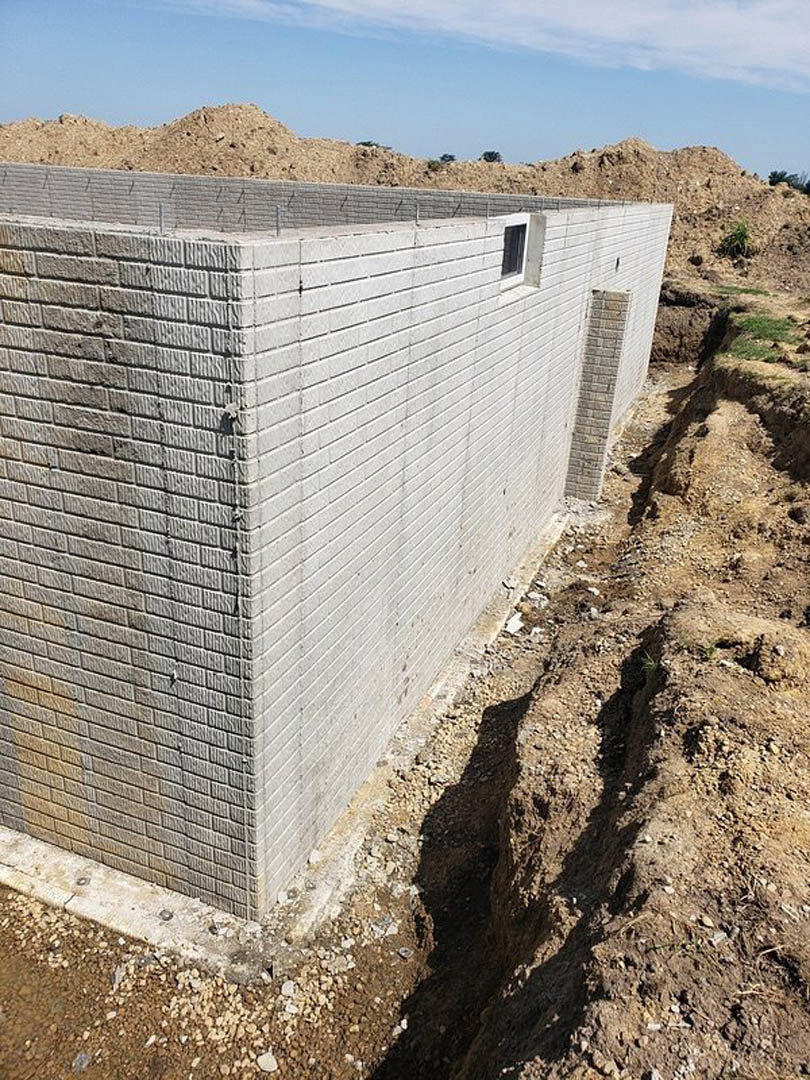 Concrete exterior wall with rectangular window, brick pillar, wire fencing, dirt excavation, patches of grass, and blue sky with scattered clouds