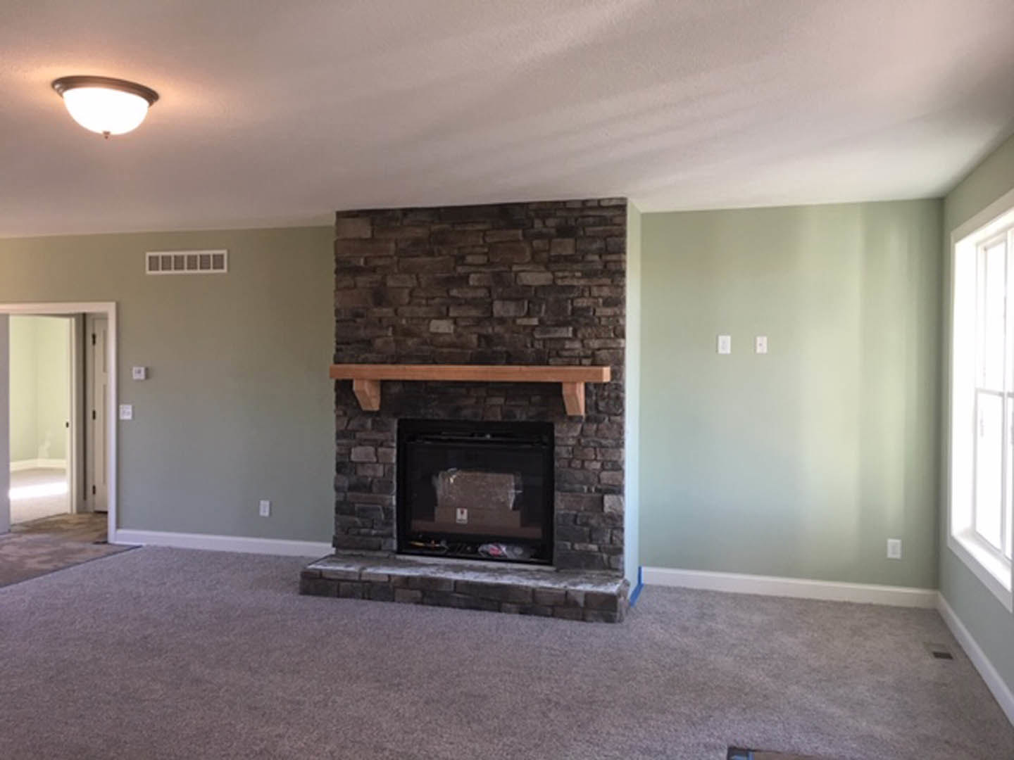 Stone fireplace with wood mantel in carpeted living room, light fixture on ceiling, white walls, and hearth with firewood box