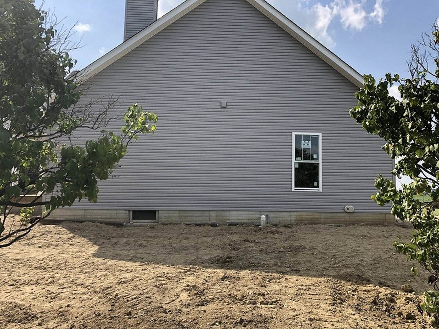 Two-story house with light siding, large windows, and a dirt yard bordered by a low wall; leafy tree in the backyard, partly cloudy sky overhead.