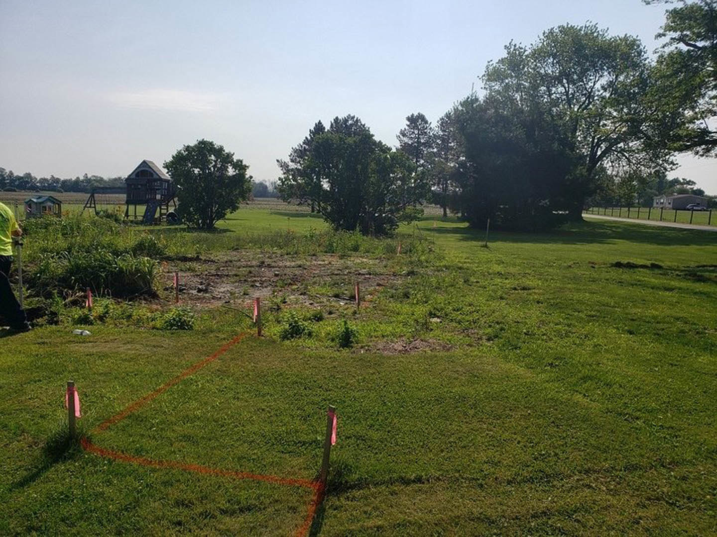 Grassy field bordered by wooden fence, scattered mature trees under partly cloudy sky