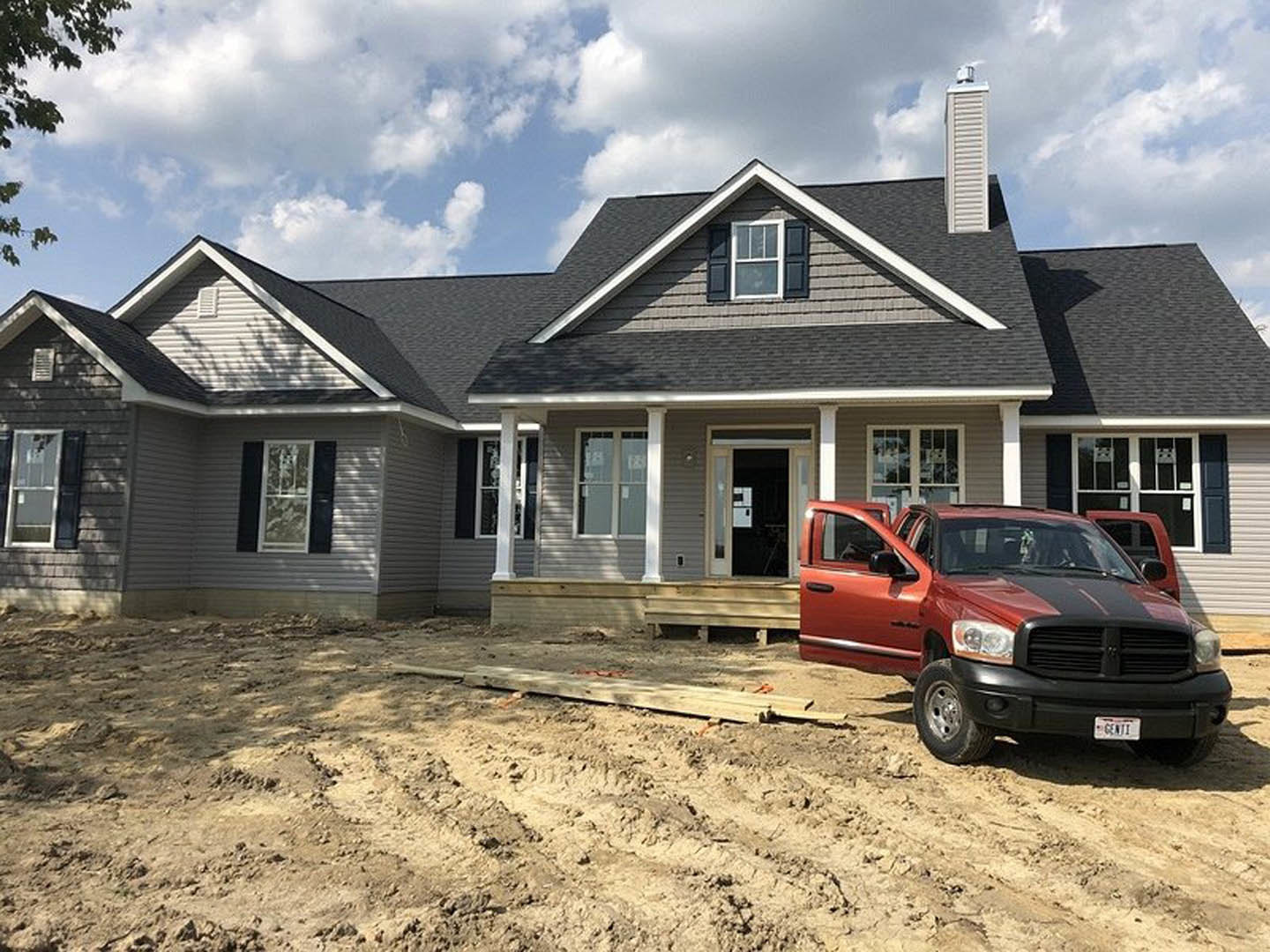 Red truck with black stripes parked on concrete driveway in front of a single-story house with frosted glass window, white door featuring a sign, and blue sky with scattered clouds