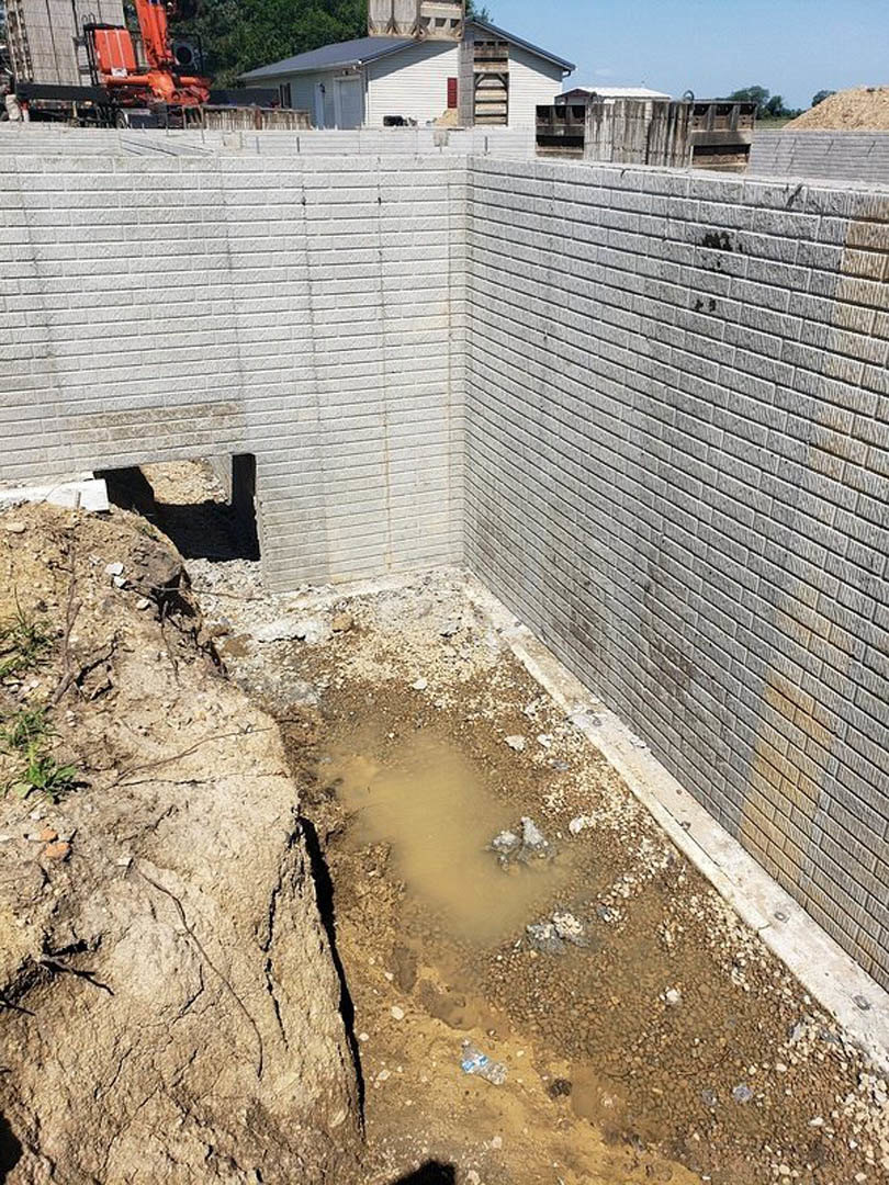 Muddy puddle on dirt ground beside red brick wall, white building with gray roof in background, blue sky with scattered clouds overhead