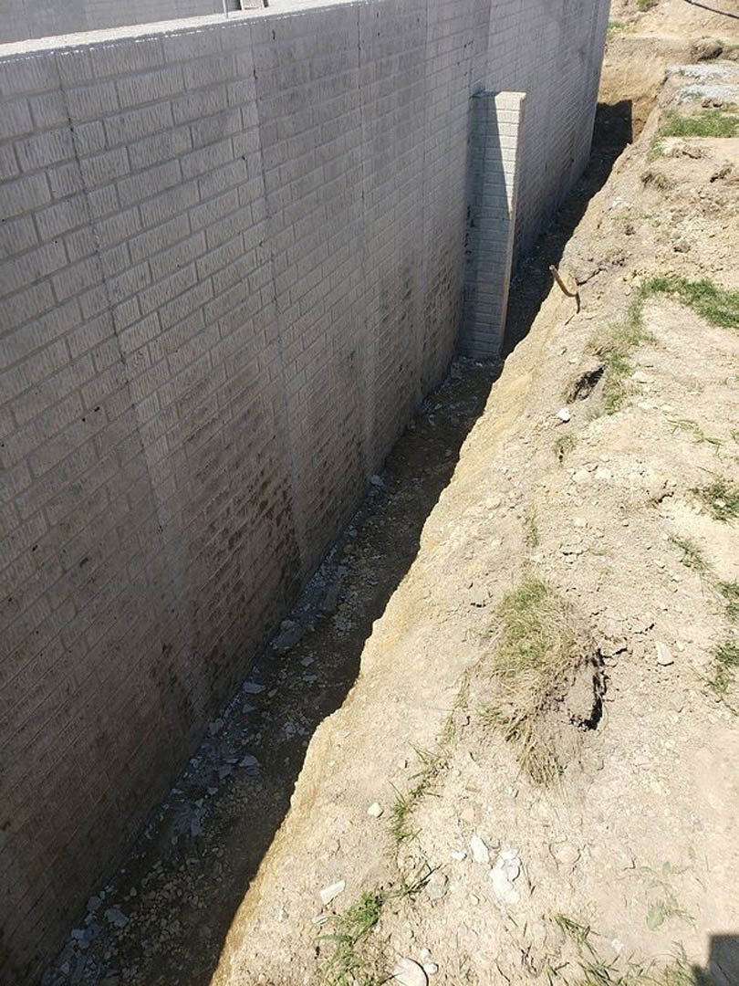 White brick exterior wall with a large hole, patch of grass at ground level, black metal pole nearby, concrete and cement surfaces visible
