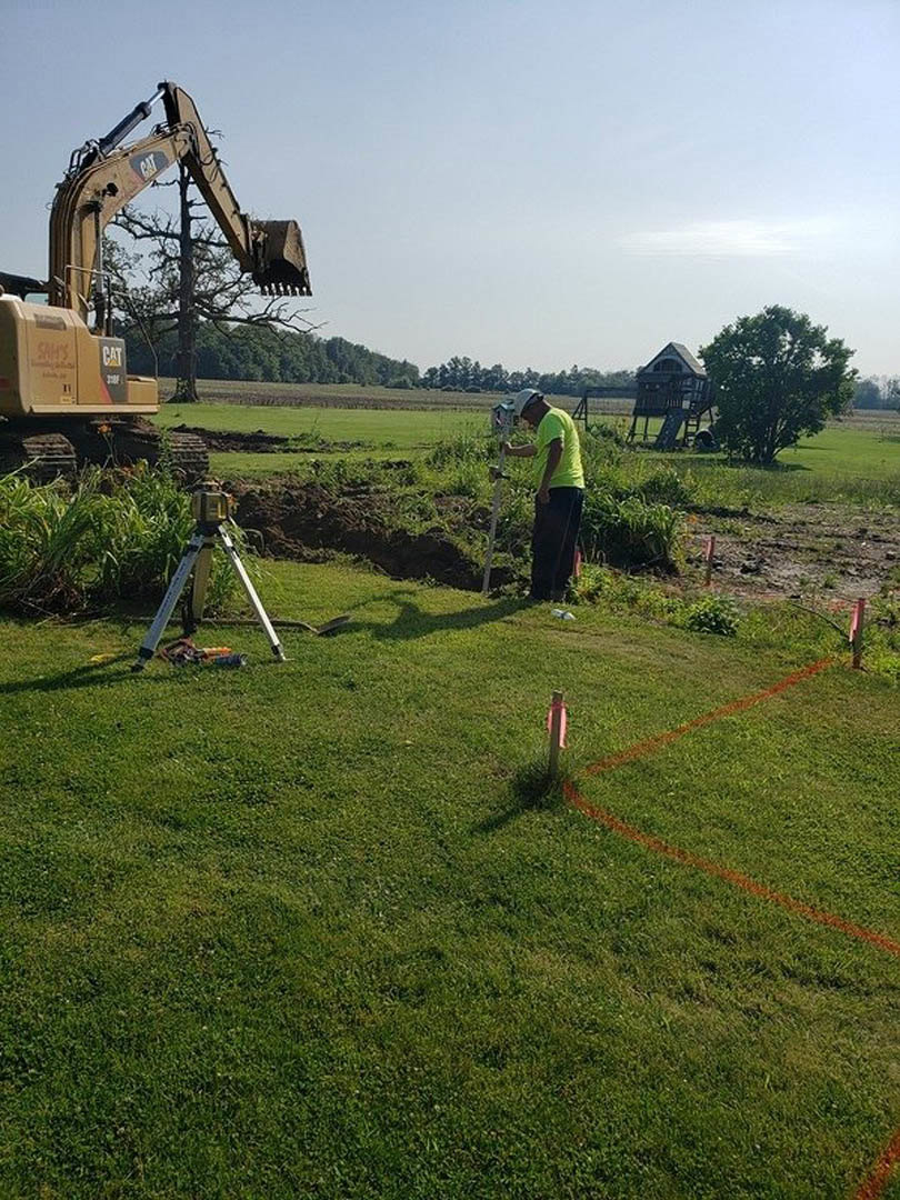 Modern custom home with white siding and large windows, surrounded by green grass and mature trees, outdoor slide attached to side of house, excavator digging near tree in open