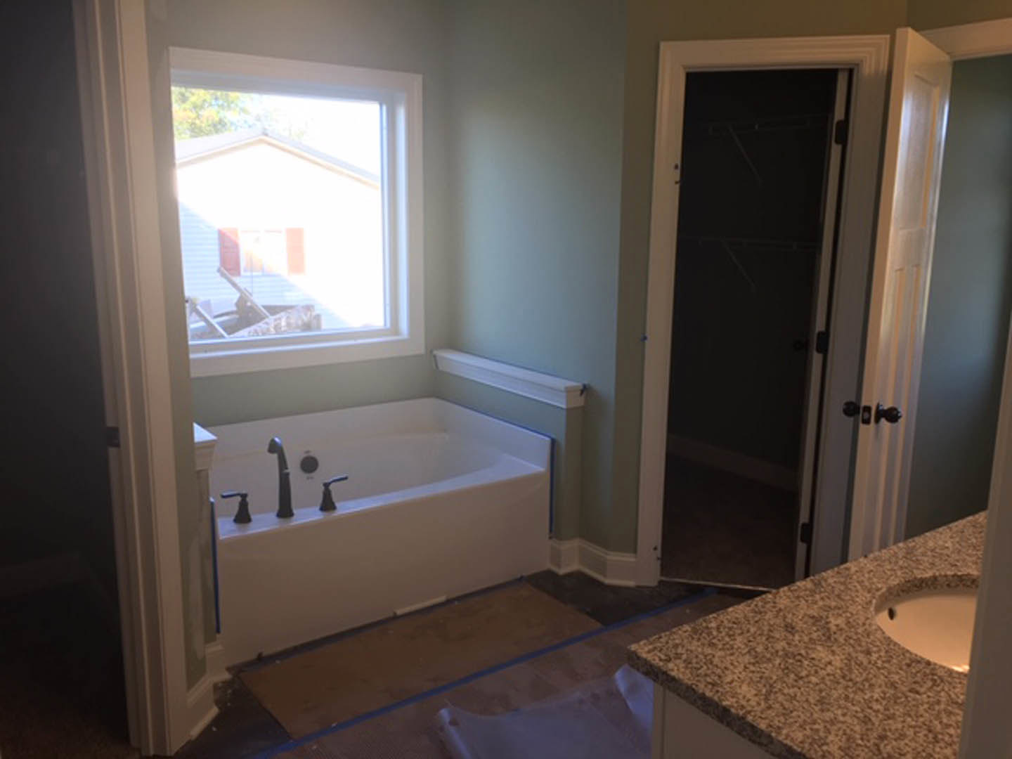 Modern bathroom featuring a freestanding bathtub with chrome faucets, white ceramic sink, tiled walls, large window overlooking a white house and tree, and dark wood door.