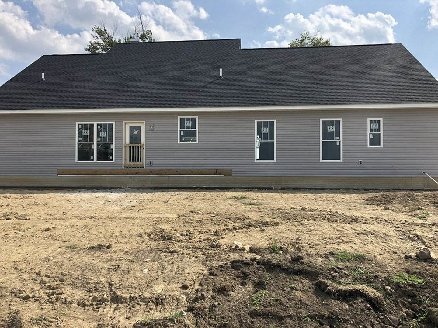 Two-story house under construction with black roof, white siding, and large windows, surrounded by dirt field and mature trees