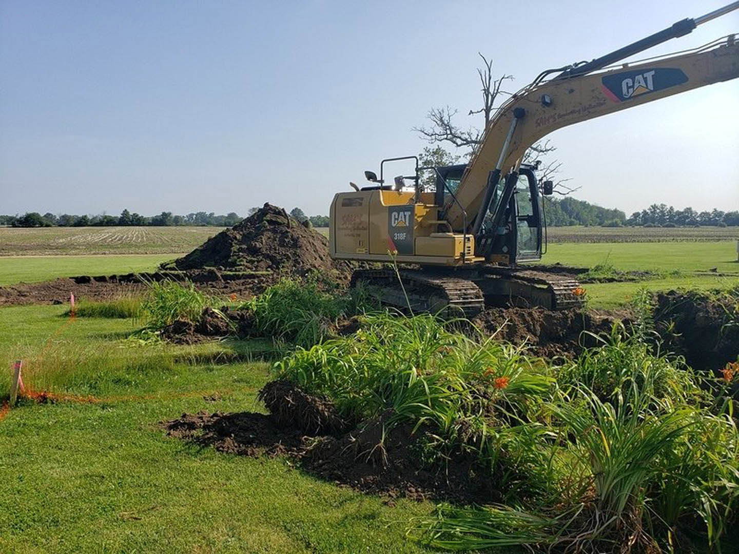 Yellow bulldozer with large arm parked on grassy field beside pile of dirt, surrounded by trees under blue sky
