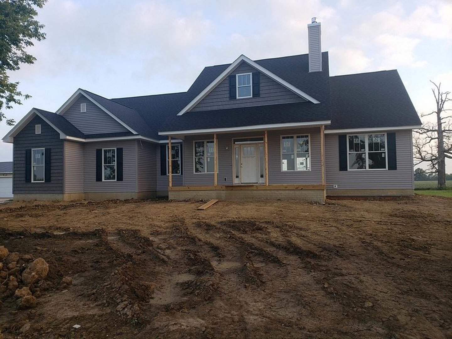 Two-story house under construction with gray siding, white porch columns, glass-paneled white front door, dirt yard with tire tracks, leafless tree, and cloudy sky