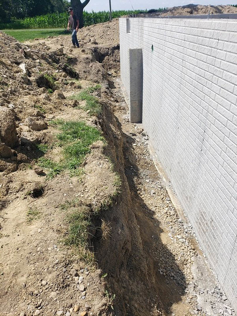 Man standing beside partially constructed brick wall with exposed trench and patch of grass in foreground, soil and trees visible outdoors.