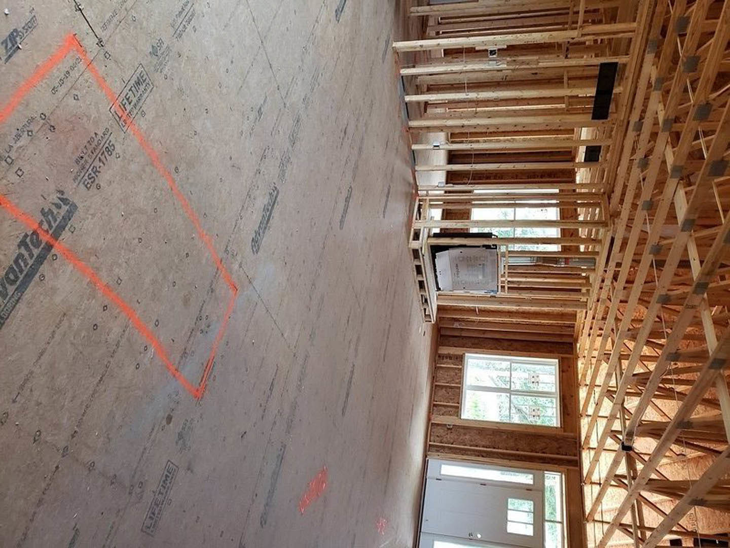Wood-framed room under construction with exposed beams, large window overlooking trees, concrete floor marked with red lines, and stack of papers on wooden shelf