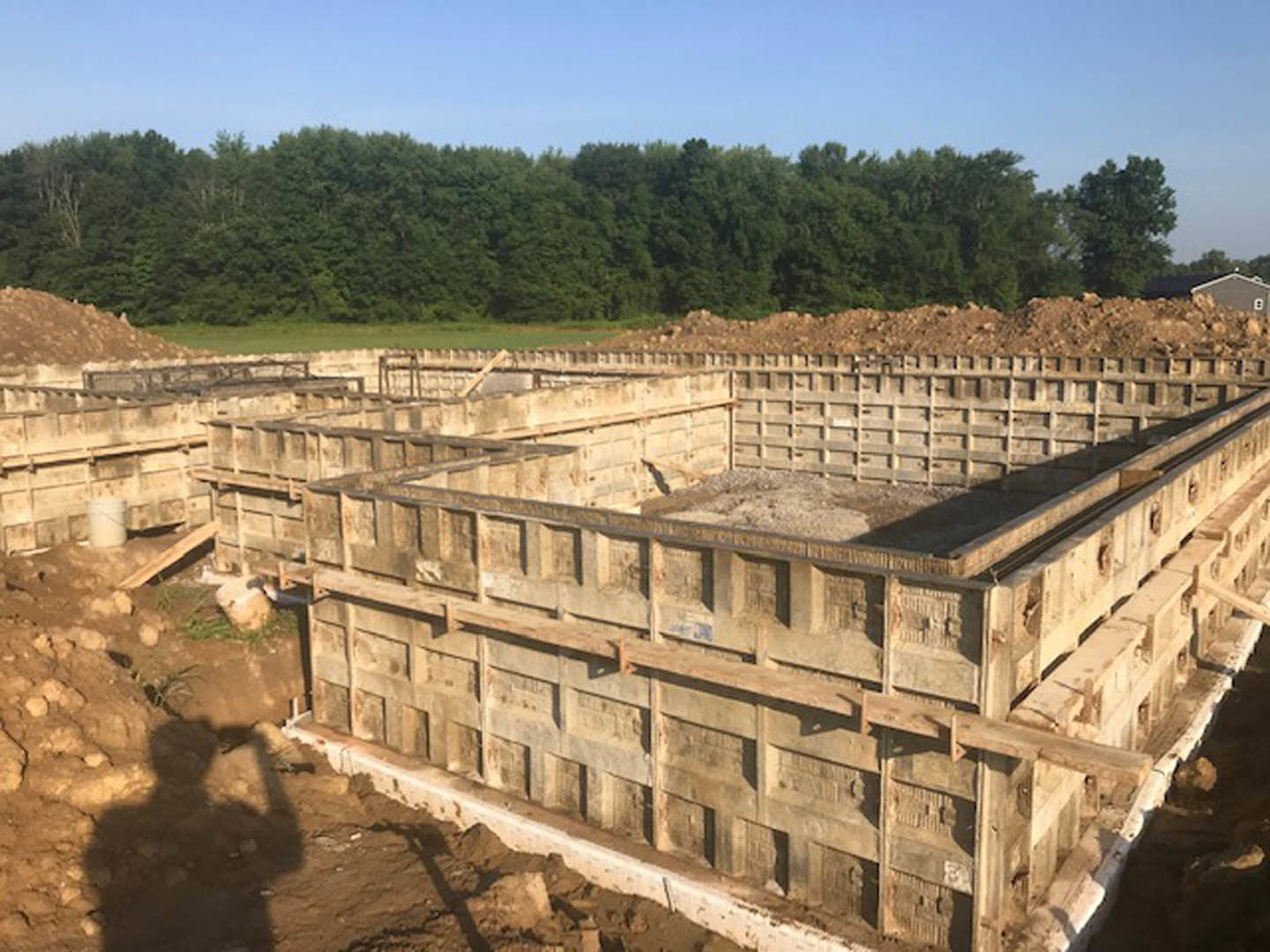Concrete foundation under construction with several concrete blocks, man standing near forested area, shadow of person holding shovel, blurry bucket and building in background