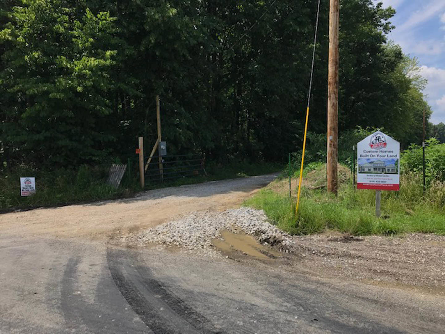 White rectangular sign with black text stands beside a dirt road bordered by trees and plants under a partly cloudy sky.