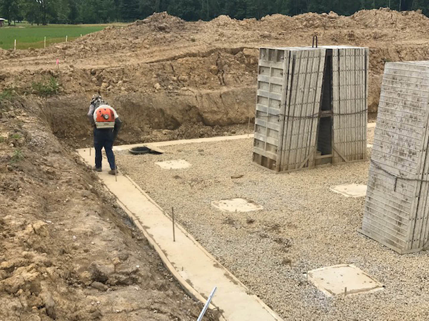 Man standing on bare soil at a home construction site with exposed foundation, surrounded by trees and outdoor equipment