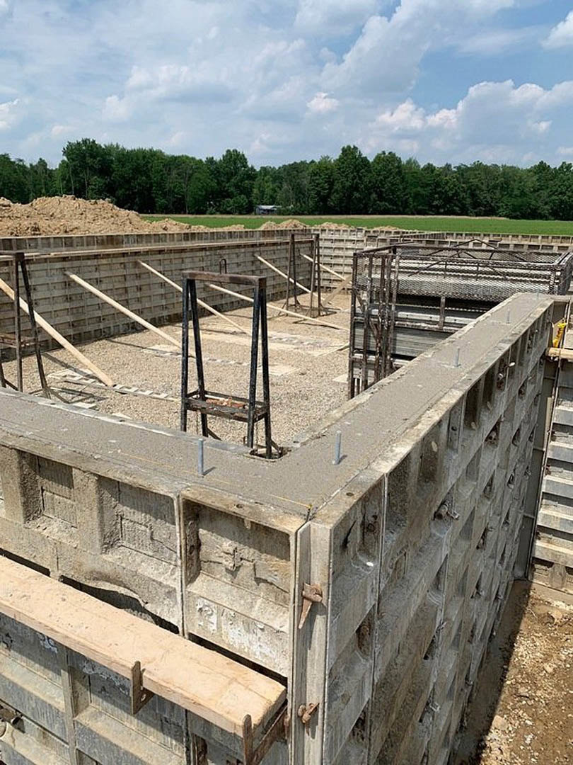 Concrete foundation with exposed metal rebar and wooden beam along wall, surrounded by grassy field and trees under cloudy sky