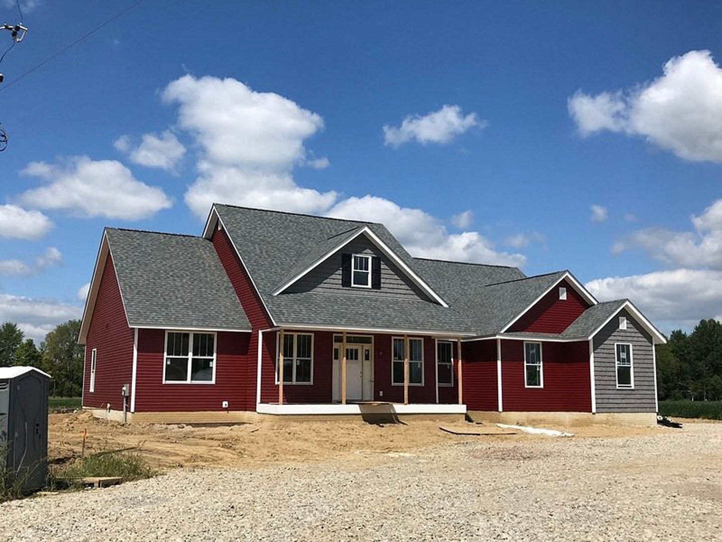 Gravel surrounds a partially built house with a red roof, white door with windows, and large windows; blue sky and clouds overhead, trees in the background